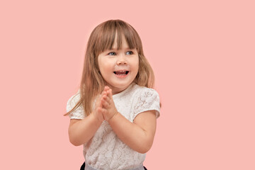 A little blonde girl in a white t-shirt smiles, looks away and claps her hands. Pink background.