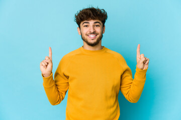 Young arab man on blue background indicates with both fore fingers up showing a blank space.