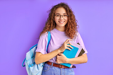 happy school girl with bag and books posing at camera isolated over purple background, cheerful...