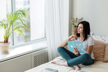 Brunette girl sitting on the bed in a bright room reading a book.