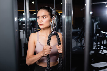 Fototapeta na wymiar athletic sportswoman looking away while exercising on cable pushdown machine on blurred foreground