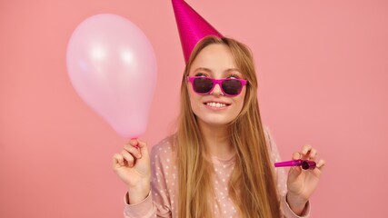 Young party woman with birthday hat holding party horn and balloon. Isolated on pink background....