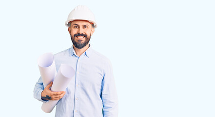 Young handsome man wearing hardhat holding paper blueprints looking positive and happy standing and smiling with a confident smile showing teeth