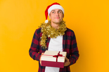 Young caucasian man with christmas hat holding a present isolated on yellow background