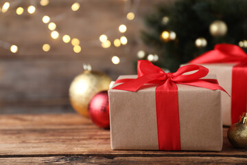 Christmas gift box with red ribbon and golden bauble on wooden table against blurred lights, space for text