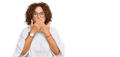 Beautiful middle age mature woman wearing casual clothes and glasses shocked covering mouth with...