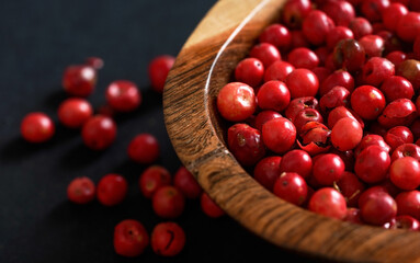 Whole pink red peppercorns in small wooden bowl, some scattered on black gray paper, closeup view from above