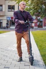 Young man in casual trousers and shirt standing next to his electric scooter, blurred city buildings background
