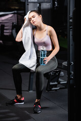 tired sportswoman wiping head with towel while sitting on training machine with sports bottle