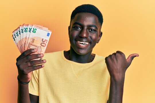 Young african american man holding 50 euro banknotes pointing thumb up to the side smiling happy with open mouth