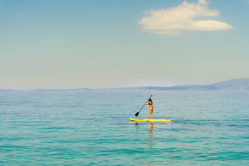 A single lonely girl on yellow paddle board or sup surf wearing red bikini paddling in the turquoise crystal clear water of mediterranean sea in Greece with single cloud  
