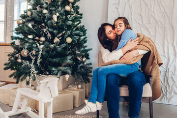 Mother and daughter hugging by Christmas tree at home. Merry Christmas and Happy New Year. Family relaxing
