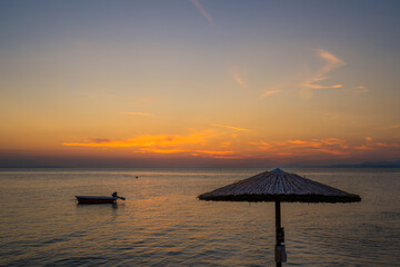 Sunset seascape with straw beach umbrella and a silhouette of a small boat anchored in a distance in Neos Marmaras, Greece 