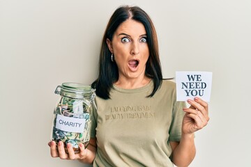 Middle age brunette woman holding charity jar with money and we need you paper afraid and shocked with surprise and amazed expression, fear and excited face.