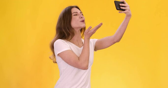 Young woman making selfie on smartphone, posing to front camera, sending blow kiss and smiling, orange background