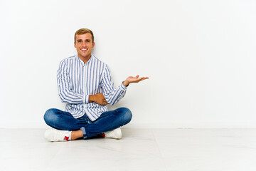 Young caucasian man sitting on the floor showing a copy space on a palm and holding another hand on...