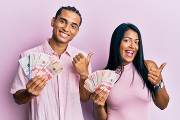 Young latin couple holding colombian pesos pointing thumb up to the side smiling happy with open...