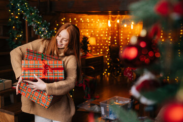 Portrait of happy smiling young woman holding box with Christmas presents wrapped in craft paper background of blurred lights. Happy lady received many presents for new year.