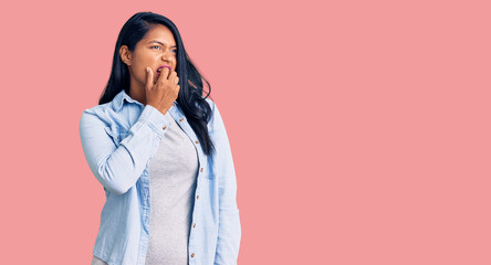 Hispanic woman with long hair wearing casual denim jacket looking stressed and nervous with hands on mouth biting nails. anxiety problem.