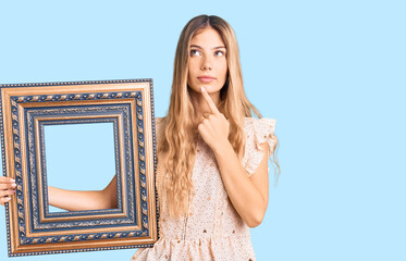 Beautiful caucasian woman with blonde hair holding empty frame serious face thinking about question...