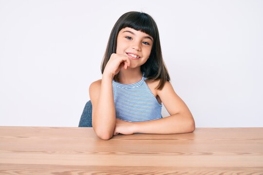 Young little girl with bang wearing casual clothes sitting on the table smiling looking confident at the camera with crossed arms and hand on chin. thinking positive.