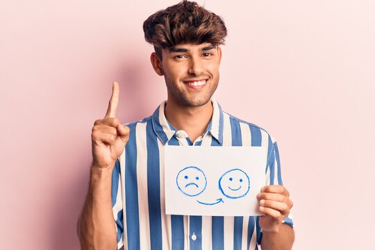 Young hispanic man holding sad to happy emotion paper smiling with an idea or question pointing finger with happy face, number one