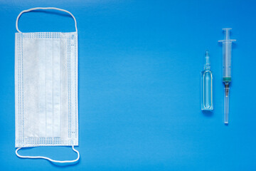 An ampoule with a vaccine, a medical mask and a syringe lie on a blue background. The concept of vaccination against coronavirus. Copy space.