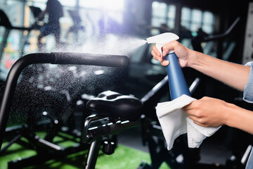 cropped view of charwoman spraying detergent while cleaning exercising machine in gym
