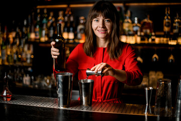 smiling woman bartender pours liquid from jigger into steel shaker cup standing on bar