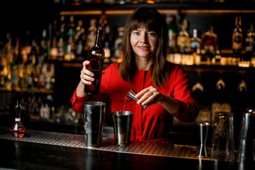 female bartender pours liquid from jigger into steel shaker cup standing on bar