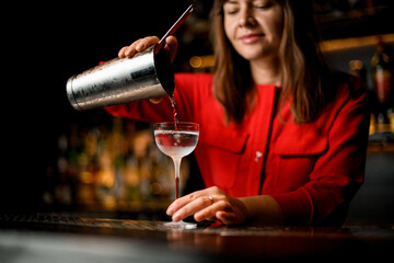 female bartender pours transparent drink from steel glass into frozen glass on bar counter