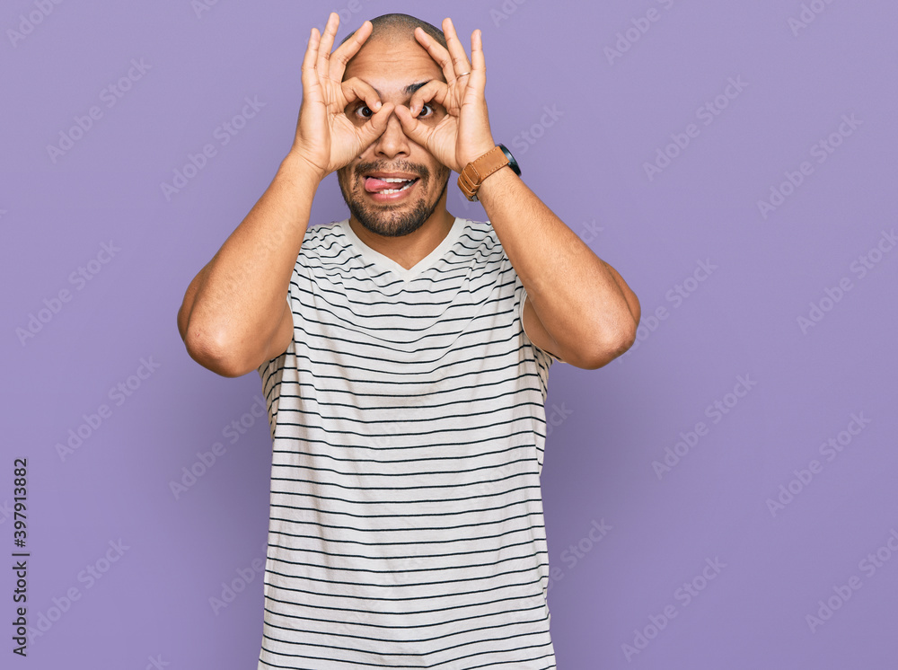 Sticker Hispanic adult man wearing casual clothes doing ok gesture like binoculars sticking tongue out, eyes looking through fingers. crazy expression.