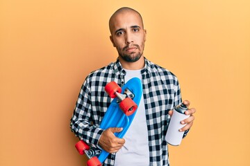 Hispanic adult skater man holding skate and graffiti spray relaxed with serious expression on face....