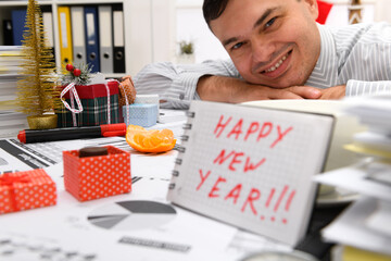 Business and holiday concept - businessman celebrating in office decorated with Christmas tree, candies and other new year decoration. Shows a notepad with happy new year text