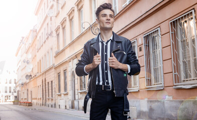 Attractive young hipster man in black jacket. fashionable walks in the city. Urban fresh portrait confident young man with hairstyle in fashion black denim jacket near wall outdoors.