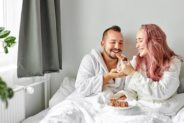 joyful couple eating eclairs in the morning lying on bed, in light bedroon, happy, smile