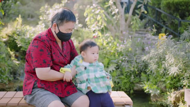 asian grandmother wearing protective fac emask takecare and look after male boy child nephew on wooden bench in garden family generation concept