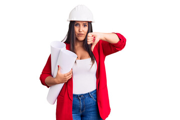 Young beautiful brunette woman wearing hardhat holding architect blueprint with angry face, negative sign showing dislike with thumbs down, rejection concept