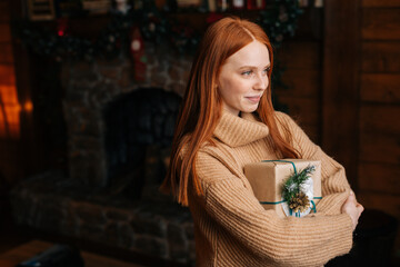 Smiling redhead young woman holding beautiful Christmas gift box while standing at cozy dark living room with festive interior. Concept of home festive atmosphere.