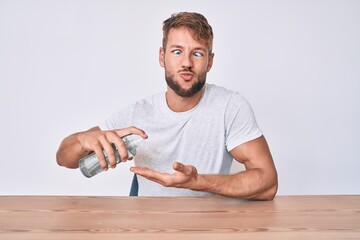 Young caucasian man using hand sanitizer gel sitting on the table making fish face with mouth and squinting eyes, crazy and comical.