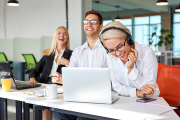 happy colleagues laughing at work place, caucasian man and women sit with laptop having fun, take a break. focus on blonde female in headphones