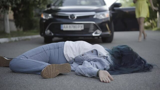 Wide shot of young woman hit by car lying on asphalt road with blurred female driver running from vehicle from background to body. Road accident on summer day outdoors.