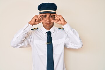 Young hispanic man wearing airplane pilot uniform trying to open eyes with fingers, sleepy and...