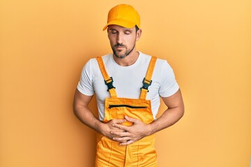 Young handsome man wearing handyman uniform over yellow background with hand on stomach because indigestion, painful illness feeling unwell. ache concept.