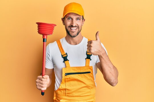 Young Handsome Man Wearing Plumber Uniform Holding Toilet Plunger Smiling Happy And Positive, Thumb Up Doing Excellent And Approval Sign