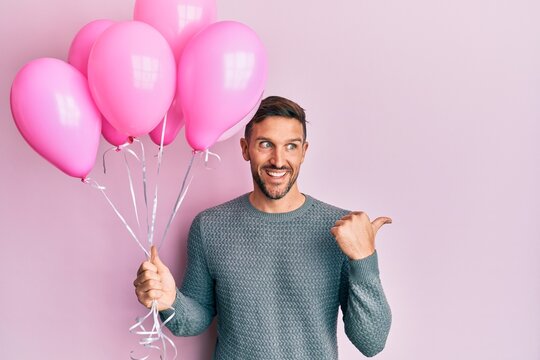 Handsome man with beard holding pink balloons pointing thumb up to the side smiling happy with open mouth