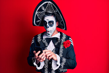 Young man wearing mexican day of the dead costume using smartphone scared and amazed with open...