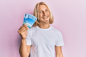 Caucasian young man with long hair holding 50000 indonesian rupiah looking positive and happy standing and smiling with a confident smile showing teeth