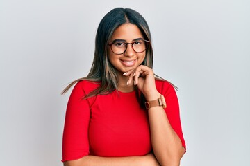 Young latin girl wearing casual clothes and glasses smiling looking confident at the camera with crossed arms and hand on chin. thinking positive.