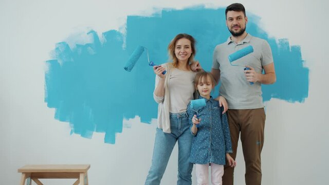 Slow motion portrait of happy family mother father and daughter holding painting rollersstanding indoors at home with painted wall in background